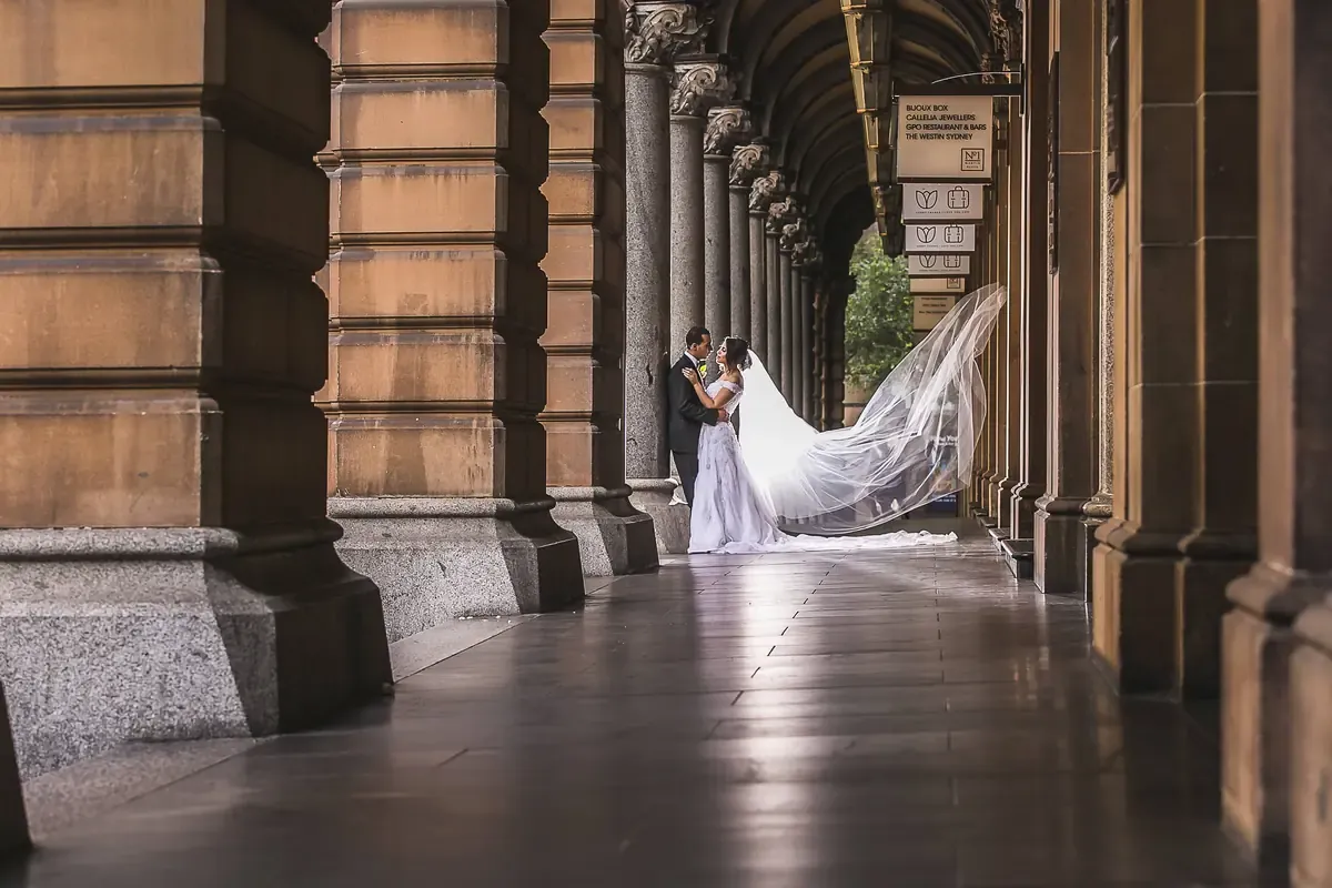 A couple standing together beneath the stone arcade at Martin Place in Sydney CBD with the veil flowing behind the bride