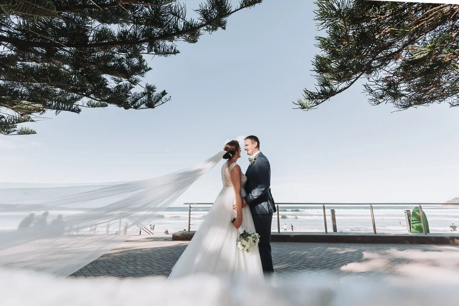 Megan and Scott standing together by the ocean at Manly Golf Club in Sydney’s Northern Beaches with the veil streaming across the shoreline