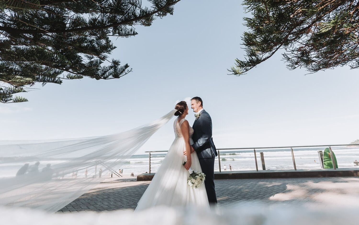 Megan and Scott standing together by the Manly shoreline beneath Norfolk pines as the veil streams across the ocean view